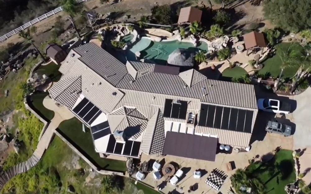 Aerial view of a large tile-roof home with solar pool heating collectors across several roof planes above a freeform pool