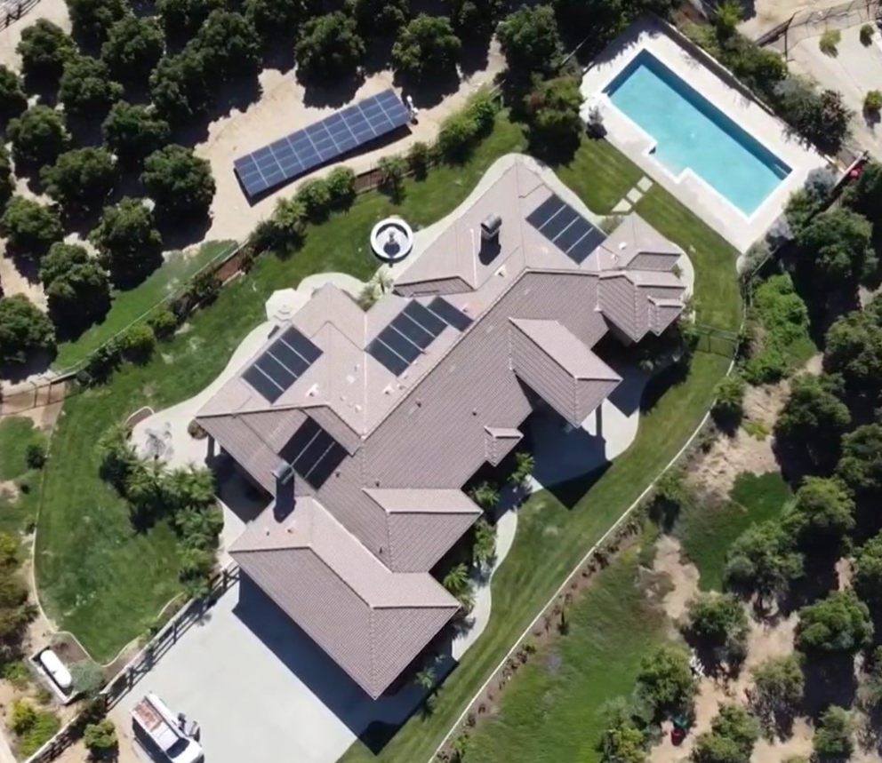 Aerial view of a home with solar collectors on the roof and an additional ground-mounted rack beyond the fence, beside a rectangular pool