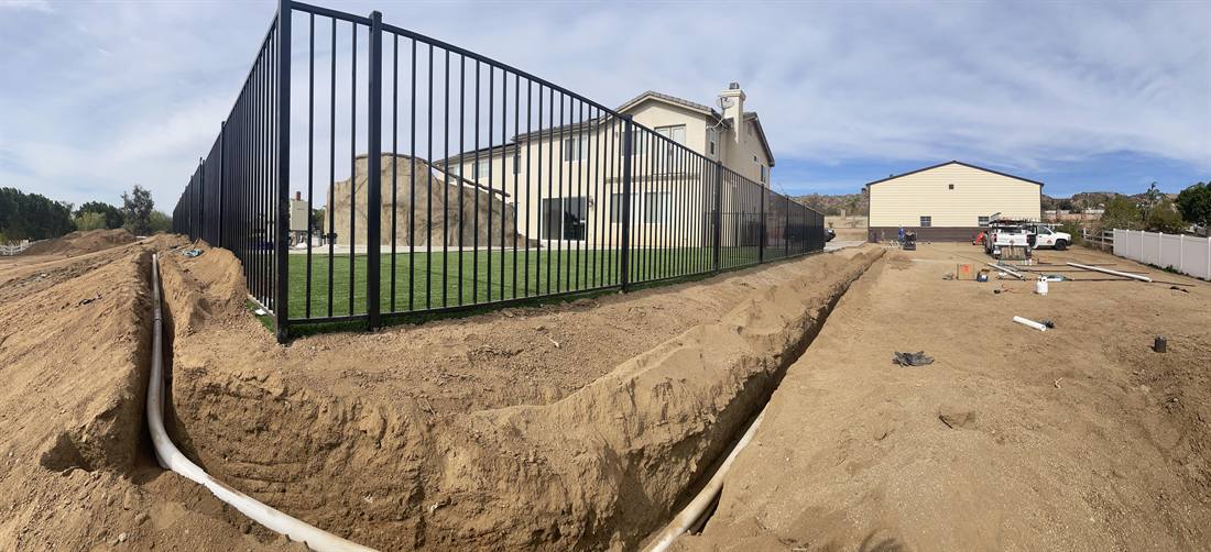 An open trench carrying white PVC solar plumbing between two homes, with a service truck on the lot