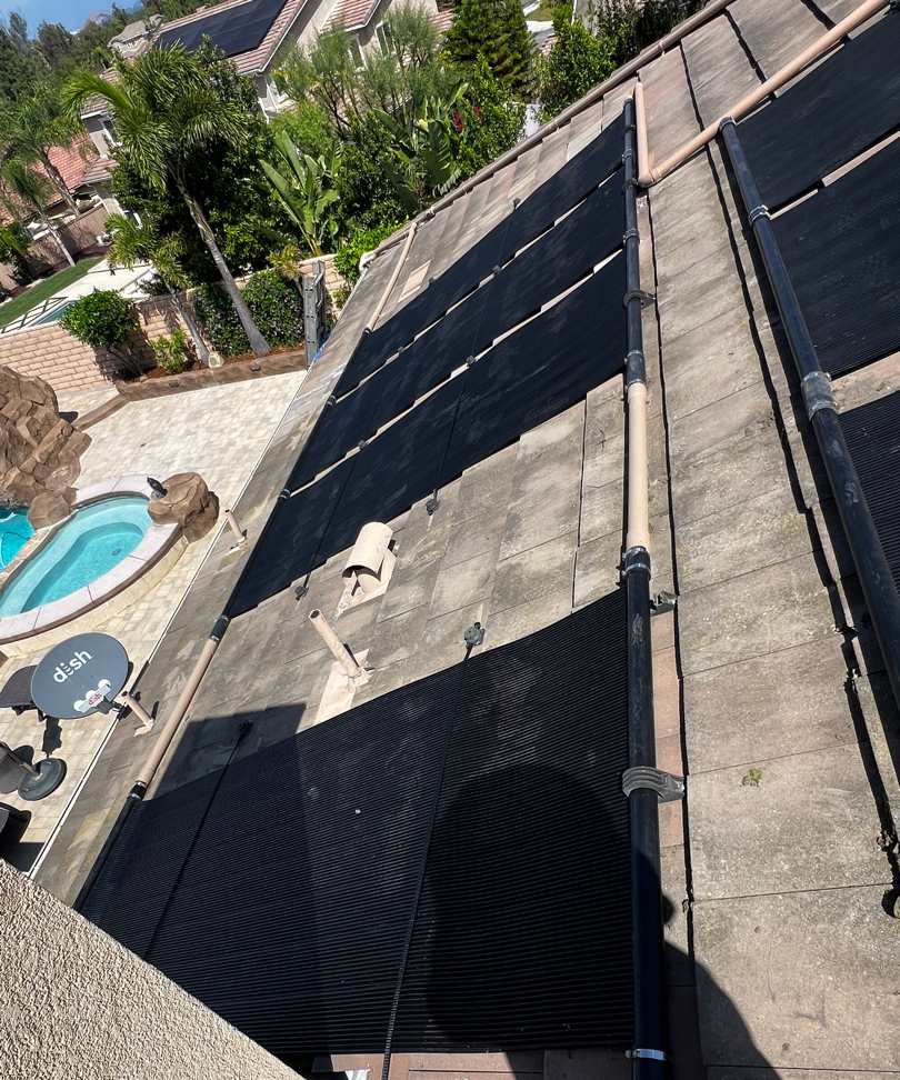 Solar pool heating collectors on a steep two-story tile roof above a backyard pool with a rock waterfall